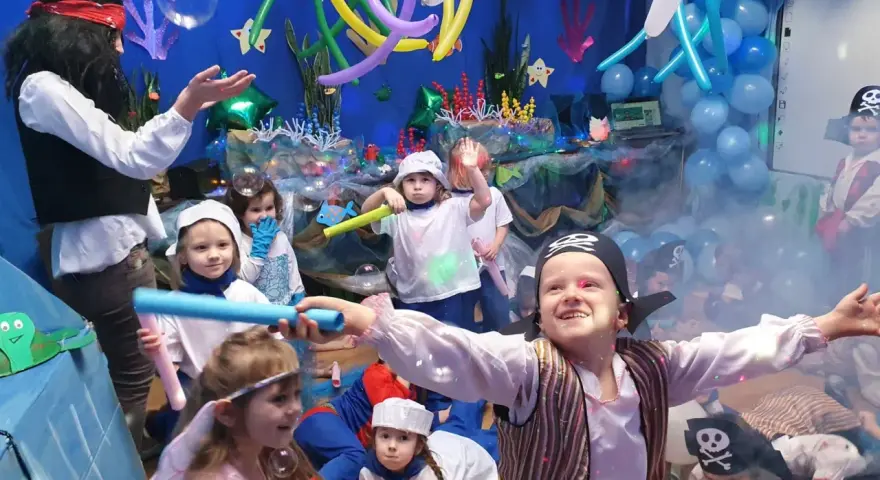 Dzieci w strojach piratów i wróżek bawią się na podwodnym balu karnawałowym. Balony i dekoracje tworzą atmosferę morskiej przygody.