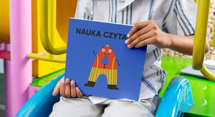Chłopiec trzyma książkę "Nauka czytania dla dzieci 6 lat" z zabawną literką "A" na okładce.
