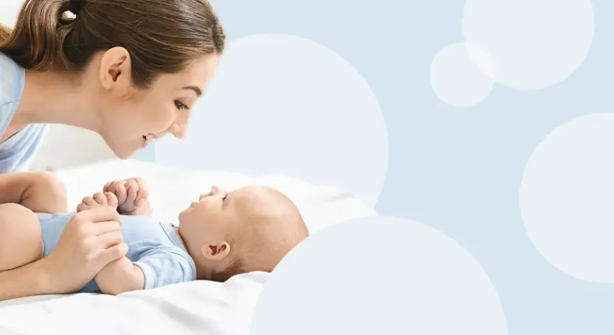 Uśmiechnięta mama pochyla się nad dzieckiem, które leży na białym tle. To magiczny moment, kiedy dziecko zaczyna mówić mama.