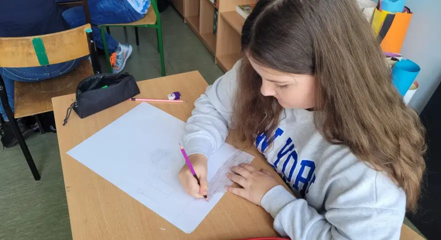 Dziewczynka rysuje w zeszycie, wykonując zadania z polskiego klasa 3. Na biurku leży czerwony piórnik.