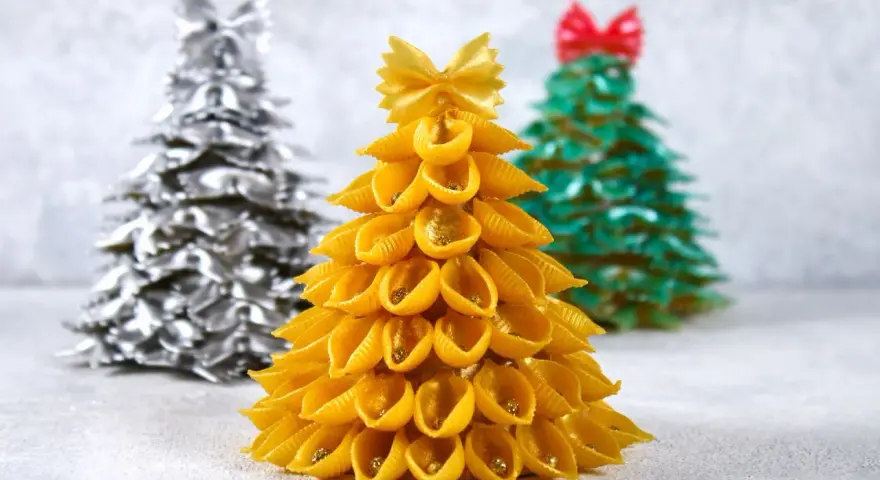 Choinka DIY dla dzieci z makaronu muszelki, ozdobiona złotym brokatem. W tle choinki z makaronu farfalle w kolorze srebrnym i zielonym.