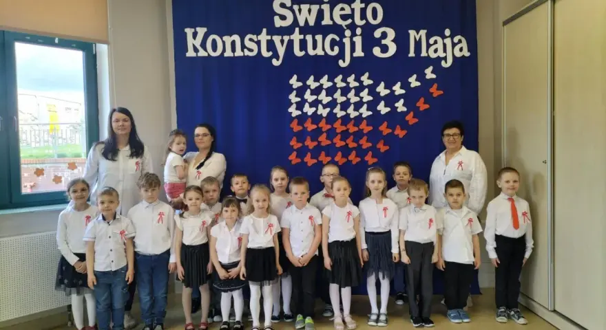 Dzieci w odświętnych strojach pozują do zdjęcia z nauczycielkami na tle niebieskiej kotary z napisem "Święto Konstytucji 3 Maja".