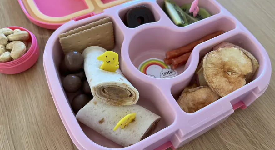 Różowy lunchbox z pysznym, zdrowym śniadaniem do szkoły: wrap z kurczakiem, suszone jabłka, kiwi, orzechy i czekoladowe kuleczki.