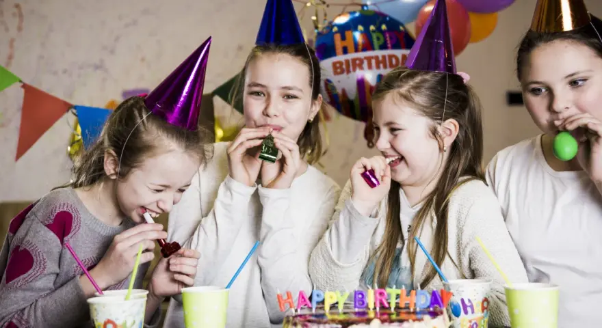 Radosne dzieci w czapkach urodzinowych dmuchają w gwizdki, świętując w domu. W tle balony i tort. Idealne animacje na urodziny w domu!