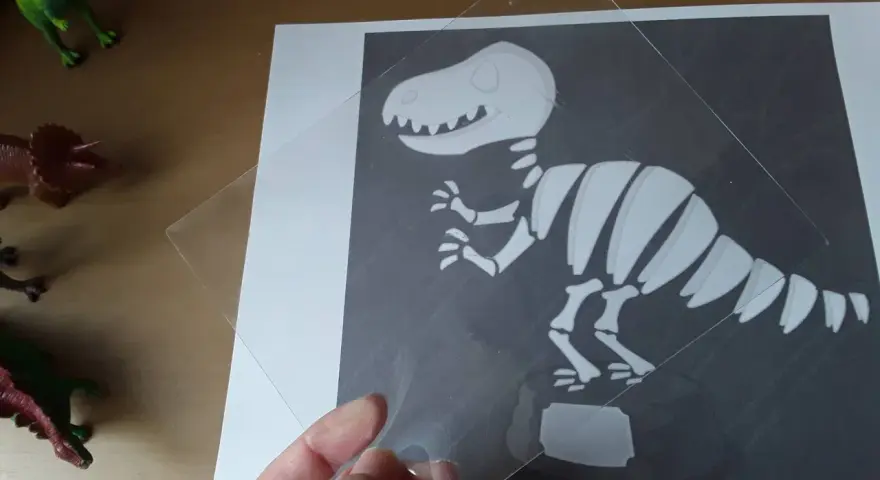 Ręka trzyma przezroczystą folię nad rysunkiem szkieletu dinozaura. To ciekawa szkielet dinozaura praca plastyczna.