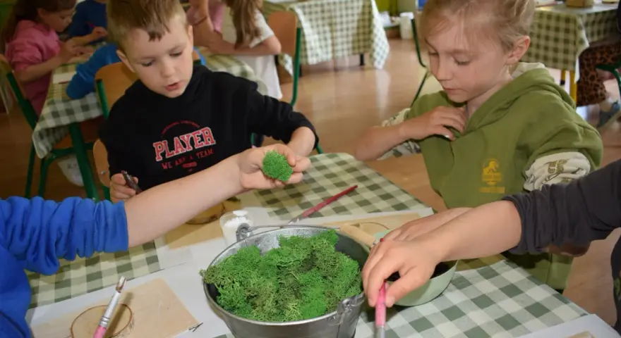Dzieci tworzą leśne dekoracje na warsztatach. W wiaderku mech, a w rękach dzieci pędzelki i drewniane krążki. Świetne pomysły na warsztaty dla dzieci!