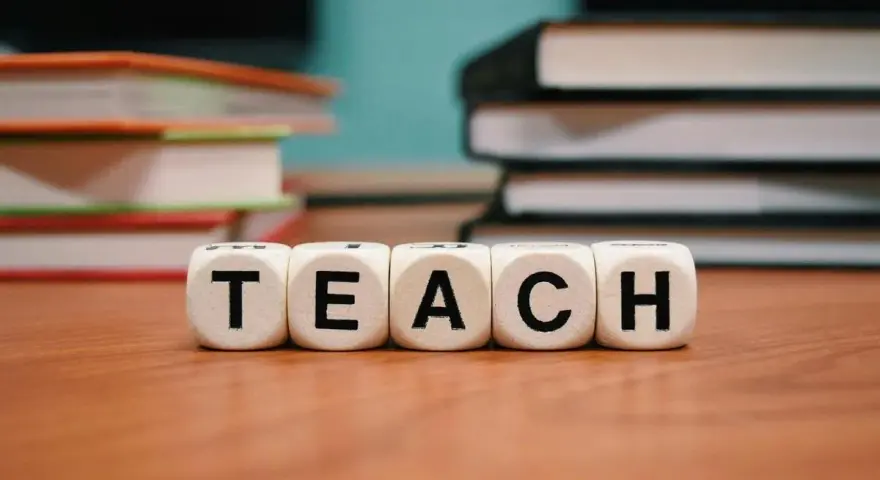 Kostki z literami tworzą słowo "TEACH". W tle stosy książek, idealne do nauki wyrazów na b dla dzieci.