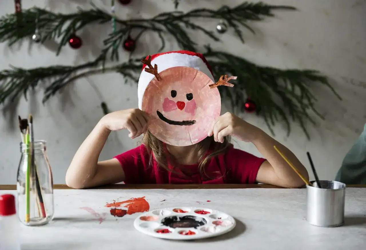 Dziecko w czapce Mikołaja tworzy świąteczne diy dla dzieci – maskę renifera z talerzyka. Na stole farby i pędzle.