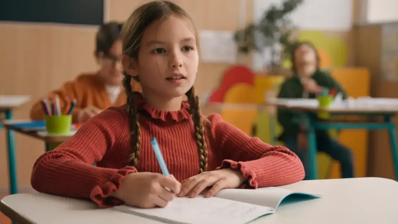 Dziewczynka z warkoczami pisze w zeszycie, tworząc opowiadanie klasa 3. W tle inni uczniowie.