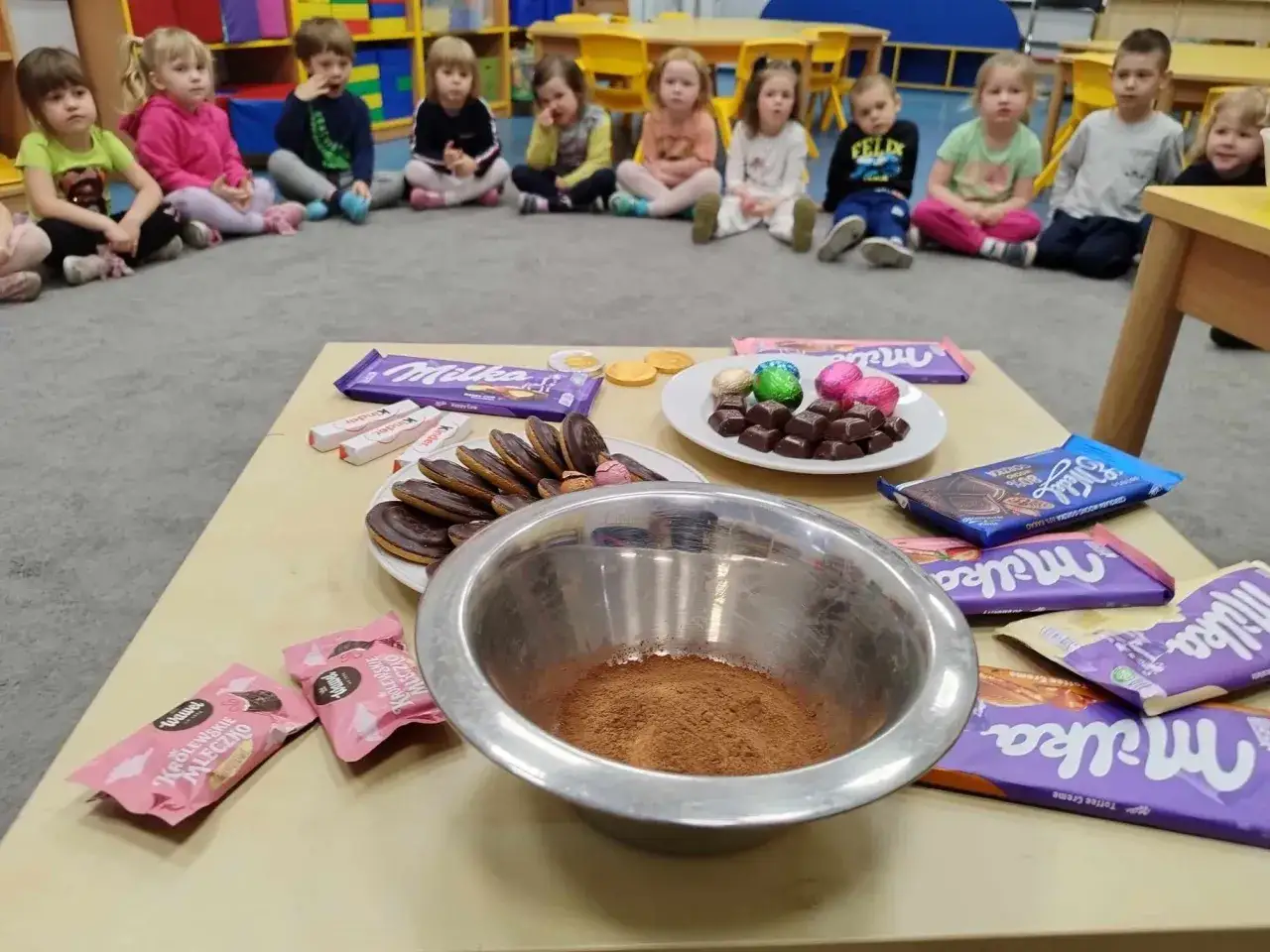 Dzieci w przedszkolu świętują dzień czekolady. Na stole czekolady Milka, Kinder, ciastka i miska z kakao.