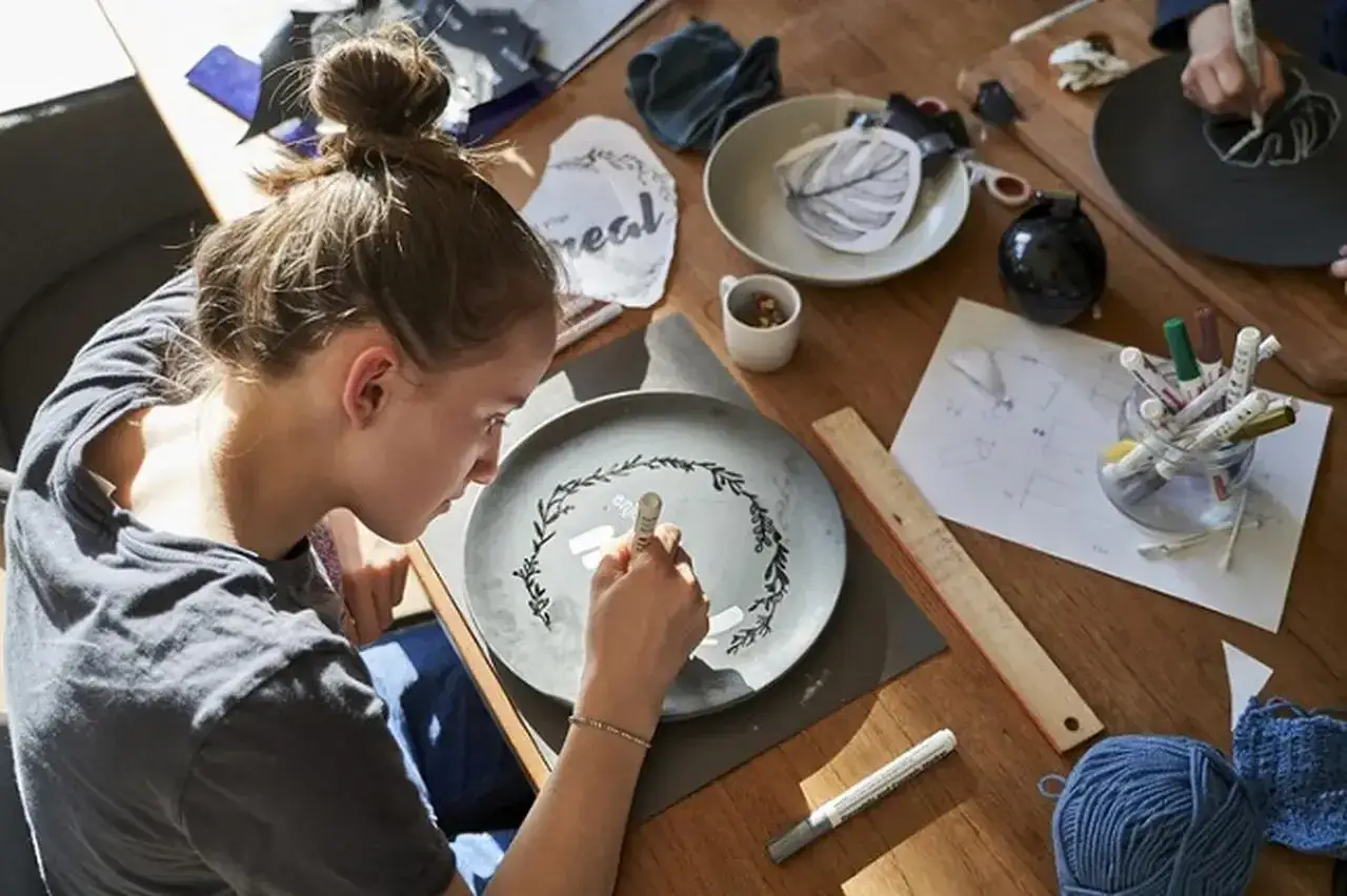Dziewczyna maluje na talerzu, tworząc wzór. Na stole materiały do rękodzieła, w tym masa porcelanowa przepis.
