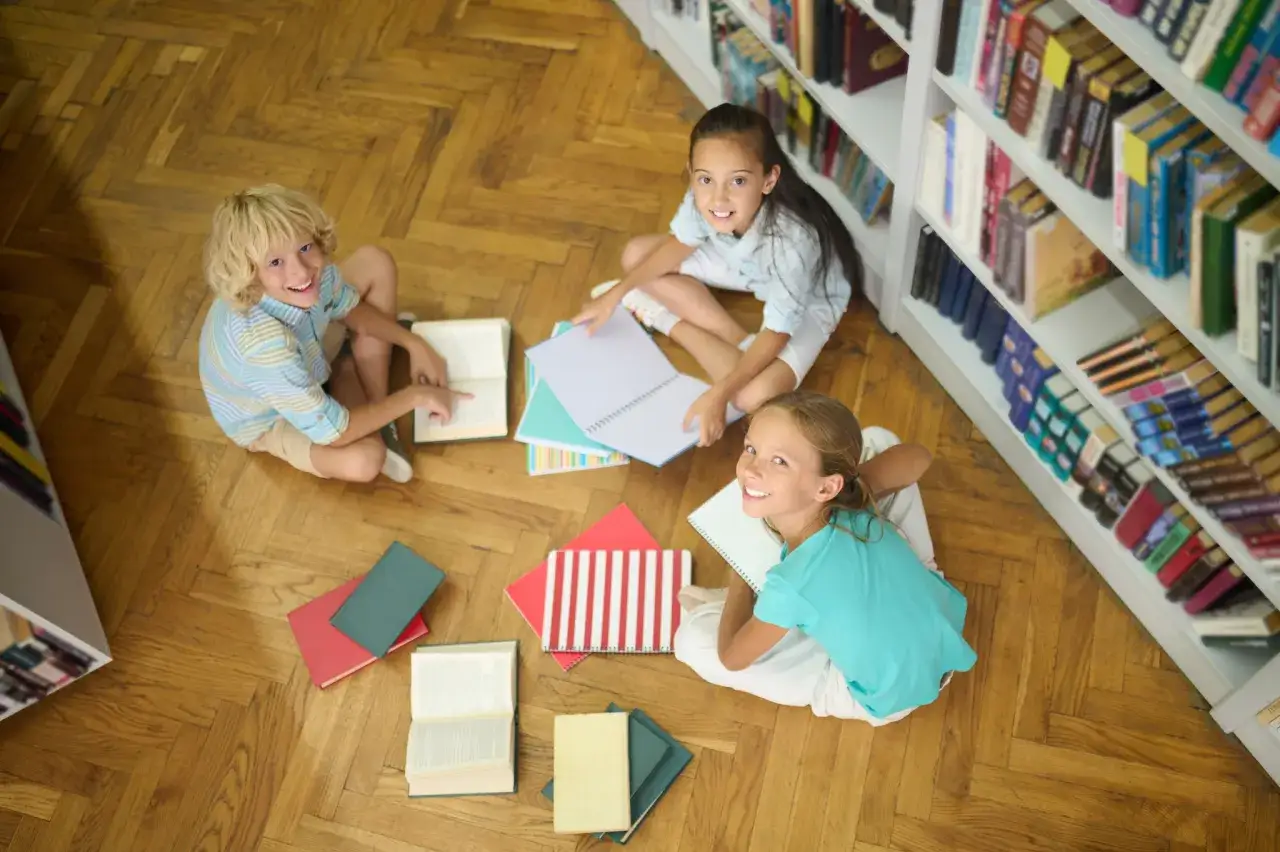 Dzieci siedzą na podłodze w bibliotece, otoczone książkami. Radośnie celebrują światowy dzień książki, odkrywając nowe historie.