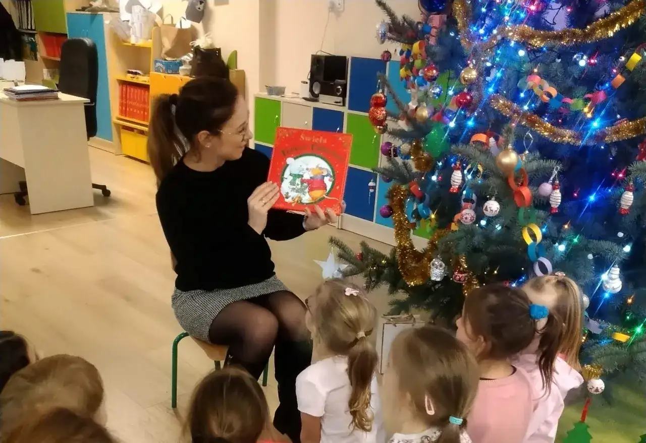 Nauczycielka czyta dzieciom książkę o świętach, pokazując jak zachęcić dziecko do czytania. Choinka w tle.
