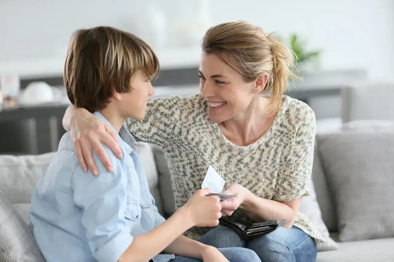 Mama daje synowi kieszonkowe. Uśmiechnięta kobieta przekazuje chłopcu pieniądze z portfela.