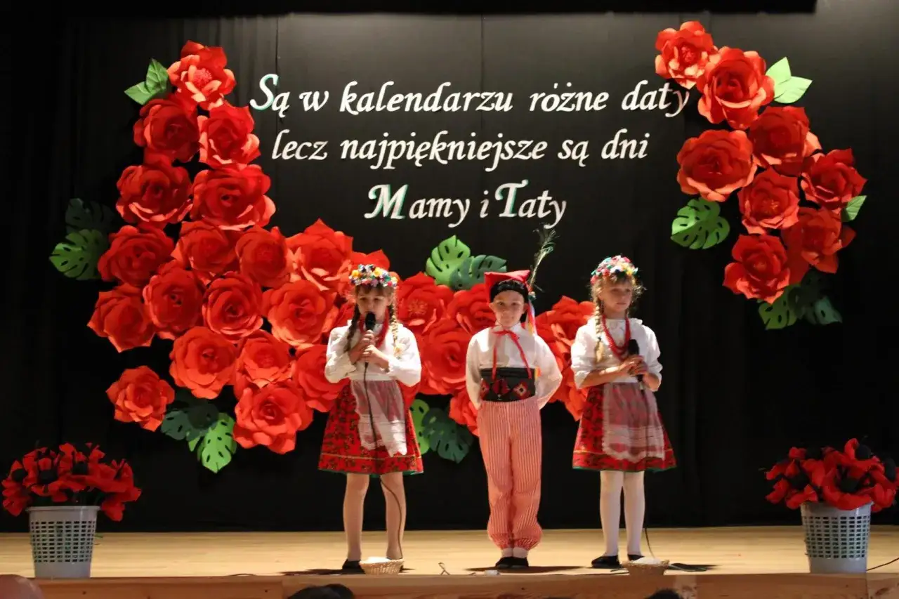 Dzieci w strojach ludowych recytują wiersz dla mamy i taty na tle czerwonych róż.