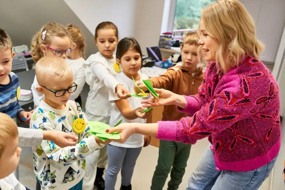 Nauczycielka z uśmiechem prowadzi zajęcia z nauki angielskiego dla 4 latka. Dzieci otrzymują zielone kartki z zadaniami.